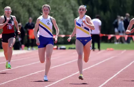Two female track athletes in a white jersey top with a yellow and a blue stripe across the front and blue bottoms, race side-by-side towards the finish line on the front stretch of the track.