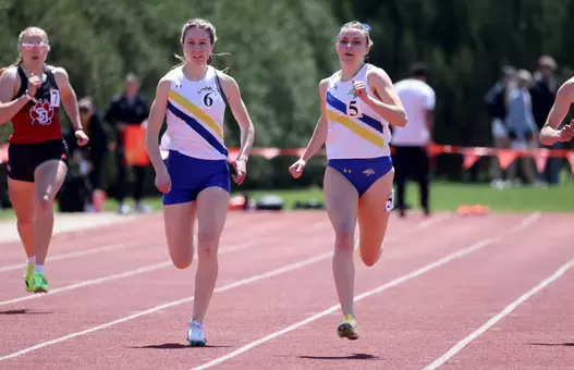 Two female track athletes in a white jersey top with a yellow and a blue stripe across the front and blue bottoms, race side-by-side towards the finish line on the front stretch of the track.