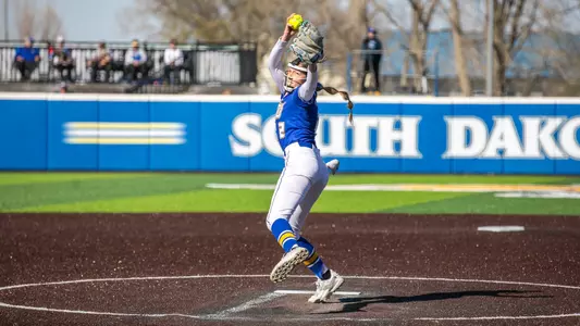 A woman in a blue softball jersey with white pants holds a softball above her head to throw a pitch