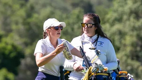Outdoor golf course scene with a blurred background of trees and greenery. In the foreground, a golfer wearing a light-colored polo, navy skirt, white cap and sunglasses stands beside a golf cart, holding a towel. Next to the golfer, a coach in a light jacket and sunglasses stands behind the cart. The two appear to be in conversation.