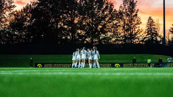 A group of women in white uniforms stand in a huddle on a soccer field during a fall evening