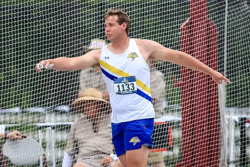 A male track athlete in a white jersey and blue shorts finishes throwing the discus and watches his attempt with his arms still outstretched.