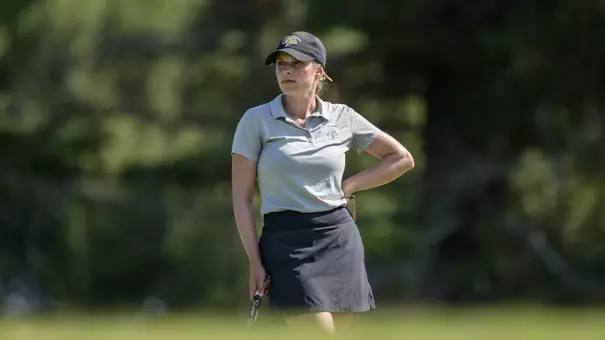 A women's golfer wearing a light colored polo and a black hat stands on a golf course, with blurred trees visible in the background
