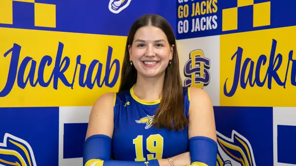 A women's volleyball player wearing a blue volleyball jersey stands with arms crossed in front of an SDSU branded backdrop