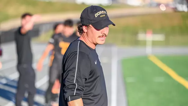 A photo of a man with a black hat and black shirt speaking with soccer players in the background