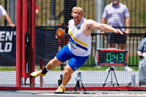 A male thrower in a white jersey and blue shorts begins to spin in the thrower's pit with an orange discus in his hand as he begins his attempt