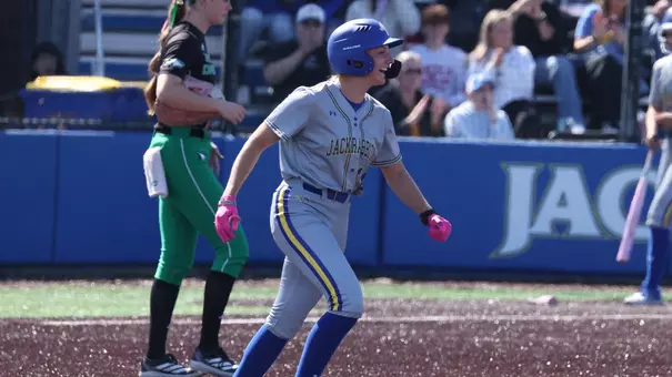 A woman in a grey softball uniform smiles with her arms raised while rounding the bases