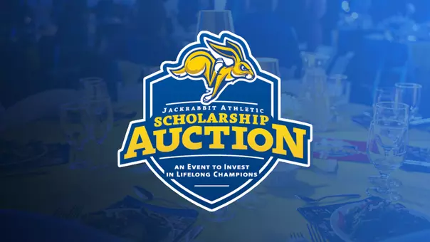 Graphic for the Jackrabbit Athletic Scholarship Auction. A large blue-and-gold shield-shaped logo with a Jackrabbit logo centered, with bold text reading “Scholarship Auction” and a subtitle that says “An Event to Invest in Lifelong Champions.” The background is a blurred banquet setting with tables, plates, glasses, and place settings, overlaid with a blue color wash.