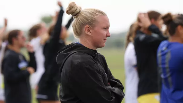 A picture of a woman in a black jacket with a group of soccer players celebrating beyond her