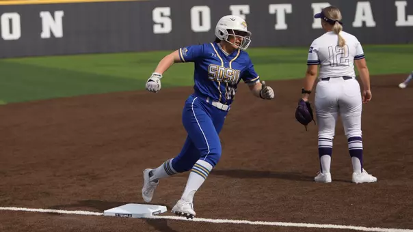 A photo of a woman in a blue softball uniform running and rounding third base