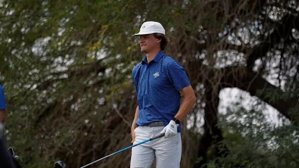 Action photo of a golfer standing on a golf course wearing a white cap, blue patterned short-sleeve polo, light-colored shorts and a golf glove, holding a club at the side. Trees and greenery fill the background.