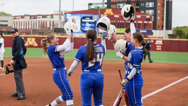 A group of softball players in blue uniforms lift their helmets at home plate while celebrating