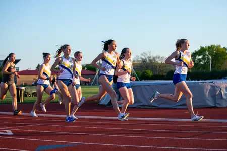 A group of five female track athletes in white jerseys and blue shorts are depicted racing around the curve of the track in their long distance running event.