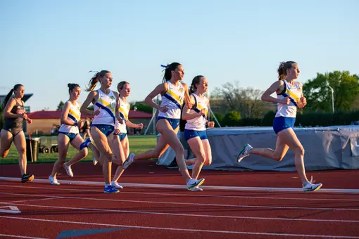 A group of five female track athletes in white jerseys and blue shorts are depicted racing around the curve of the track in their long distance running event.