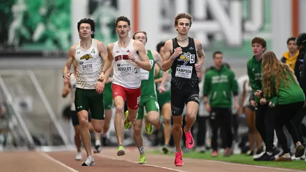 A male track athlete wearing a black uniform is depicted running head on down the inside of the track with two competitors, one in white and red, one in white and green, racing beside him.
