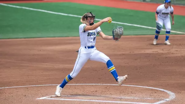 A woman in a white softball uniform lifts a softball in the air and gathers as she prepares to pitch it towards home plate
