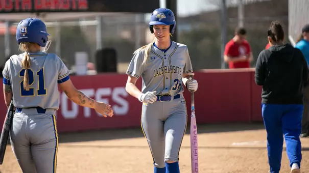 A woman in a grey softball uniform with a blue helmet fives with a teammate while holding a bat and walks back to the dugout