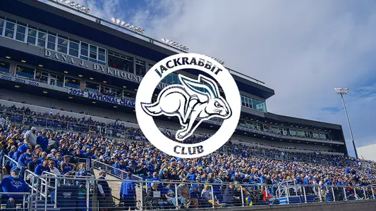 A large outdoor stadium filled with a dense crowd of spectators seated in tiered stands. The majority of people are wearing blue and yellow clothing. Centered over the image is a circular graphic overlay with a white background, displaying a stylized rabbit and the words “JACKRABBIT CLUB” surrounding it.