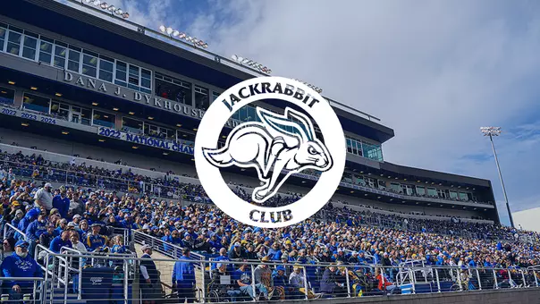 A large outdoor stadium filled with a dense crowd of spectators seated in tiered stands. The majority of people are wearing blue and yellow clothing. Centered over the image is a circular graphic overlay with a white background, displaying a stylized rabbit and the words “JACKRABBIT CLUB” surrounding it.
