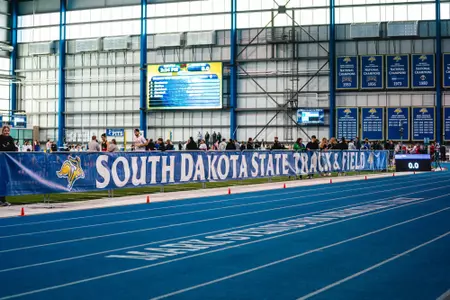 A banner runs across barriers separating the track front stretch from the infield. The banner reads, "South Dakota State Track and Field."