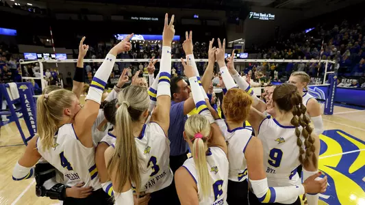 A volleyball team in white jerseys surrounds a coach in blue, while holding two fingers in the air.