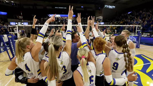 A volleyball team in white jerseys surrounds a coach in blue, while holding two fingers in the air.