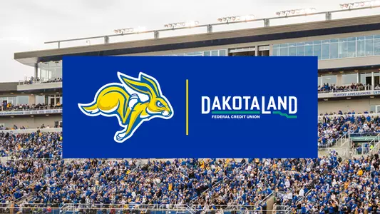 Dakota Land Federal Credit Union sponsorship graphic displayed over a football stadium crowd. A blue rectangular banner centered on the image features the South Dakota State Jackrabbits logo on the left and the Dakota Land Federal Credit Union logo on the right, separated by a vertical gold line. In the background, a packed stadium with spectators in blue and white fills the stands beneath a modern grandstand.