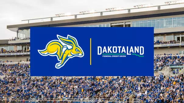 Dakota Land Federal Credit Union sponsorship graphic displayed over a football stadium crowd. A blue rectangular banner centered on the image features the South Dakota State Jackrabbits logo on the left and the Dakota Land Federal Credit Union logo on the right, separated by a vertical gold line. In the background, a packed stadium with spectators in blue and white fills the stands beneath a modern grandstand.
