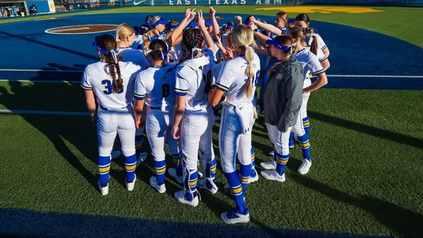 A group of softball players in white uniforms huddle on the field with their hands up in the middle of the group