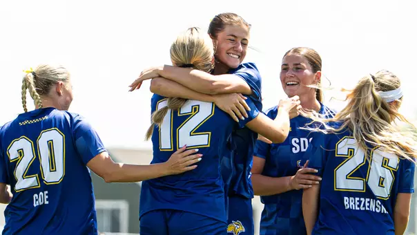 Two soccer players in blue uniforms jump into a hug in celebration while surrounded by several other teammates