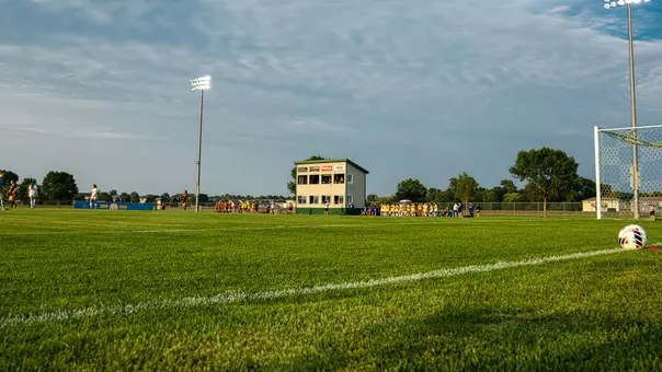 An image of a soccer field with a press box on field level and teams in huddles in the distance