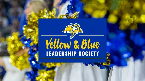 Graphic announcing the Yellow & Blue Leadership Society. A blue rectangular banner is centered over a background of blue and yellow pom-poms at a sporting event. The South Dakota State University Jackrabbit logo appears at the top of the banner above the text “Yellow & Blue” in yellow script and “LEADERSHIP SOCIETY” in white capital letters. The overall color scheme is blue, yellow, and white.