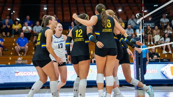 A group of volleyball players in black uniforms gather in a huddle after a point.