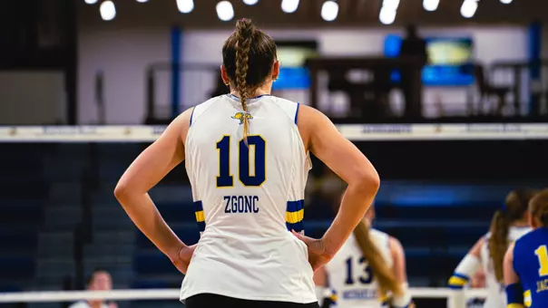 A volleyball player in a white uniform stands with her hands on her hips.