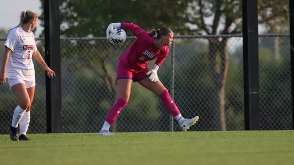 A woman in a yellow soccer uniform bends down and lifts her arm up with the ball as she prepares to roll it over to a teammate. An opponent in a white uniform runs near her.