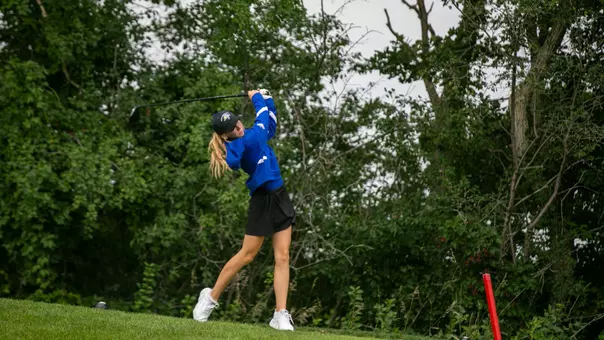 A golfer in a blue pullover and black skirt hits with a driver.