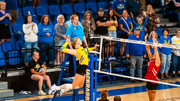 Hannah Klein in a yellow jersey, jumps to hit a ball while a player in red attempts to block.