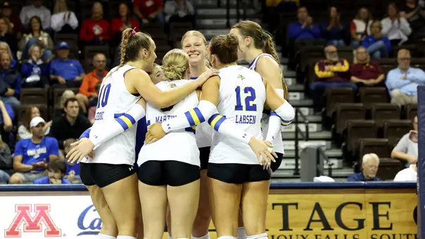 A group of players in white huddles after a point during a volleyball match.