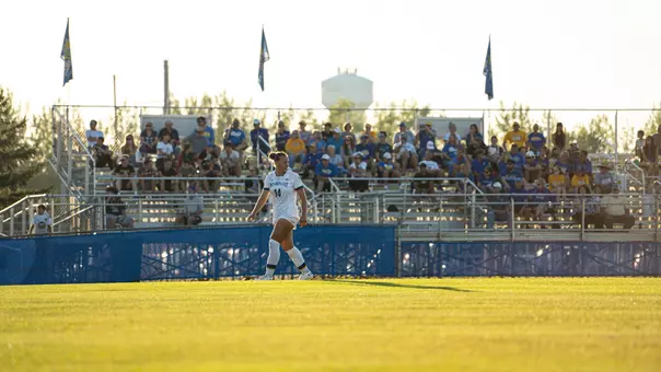 An SDSU soccer player stands on a soccer field with a set of fans sitting in a grandstand in the distance