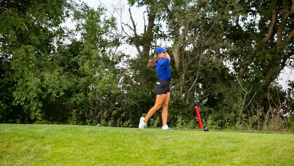 A golfer in a blue polo and cap swings a driver on a tee pad.