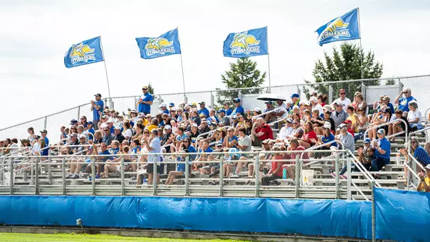 A group of fans sit in a seating section while viewing a soccer game. Flags with a Jackrabbit logo wave above the grandstand with blue wind screens covering the front-facing fence.