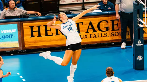 A volleyball player in a white uniform jumps to strike a ball.