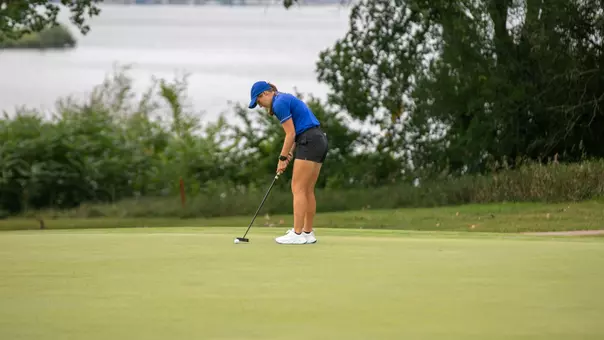 A golfer in a blue polo and cap lines up a putt on a putting green.