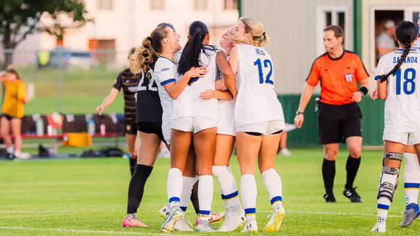 A group of women's soccer players in white uniforms celebrate together after a goal is scored