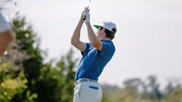 Action photo of Drew Teeter during a swing follow-through on a golf course. Teeter wears a blue patterned polo shirt, white pants, a white cap, and a golf glove, with the club raised overhead after striking the ball. Trees and greenery are visible in the blurred background under a bright sky.