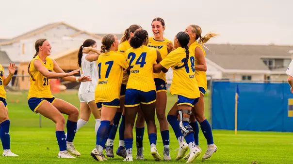 Players in yellow uniforms group up in celebration of a goal as a teammate smiles after the score