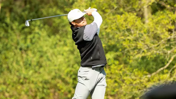 Ty Adkins follows through on a tee shot during competition. The golfer wears a white Jackrabbit cap, black vest, striped long-sleeve shirt, and gray pants. A golf club extends behind the golfer at the end of the swing, with trees and green foliage blurred in the background on a sunny day.