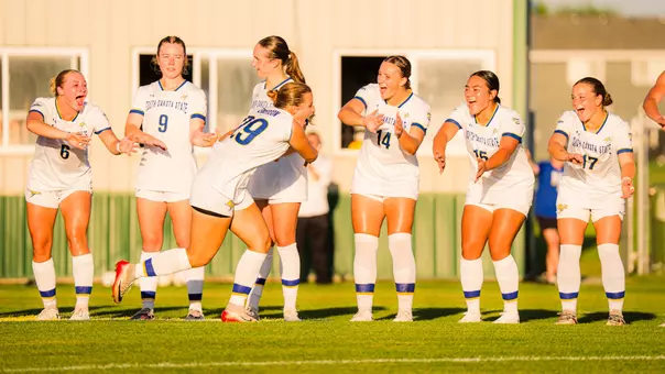 Women's soccer players in white uniforms line up and high five as they get ready to start a match, with one player running through the line