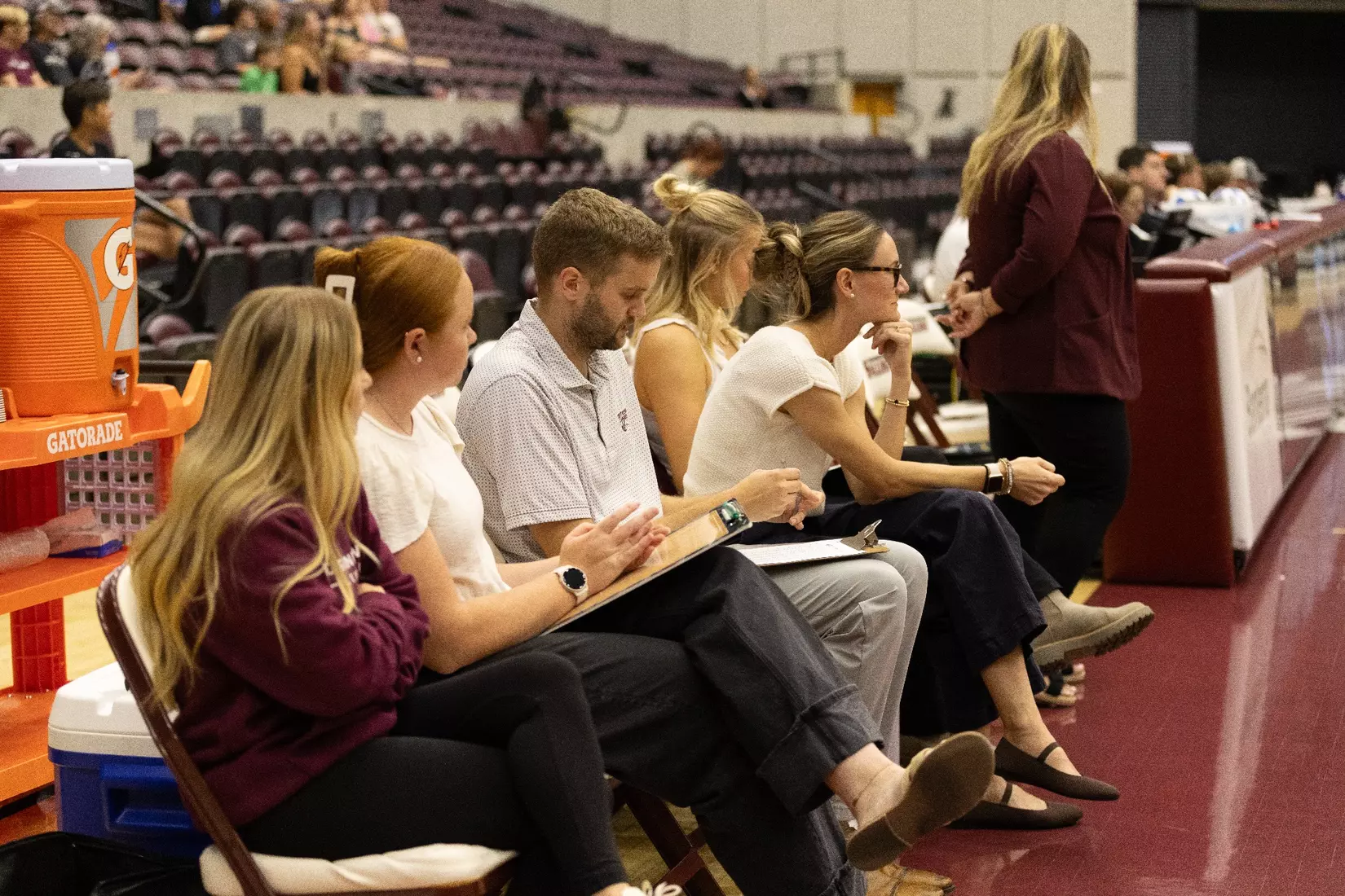 The coaching staff sits on the bench and watches as the opposing team is announced.