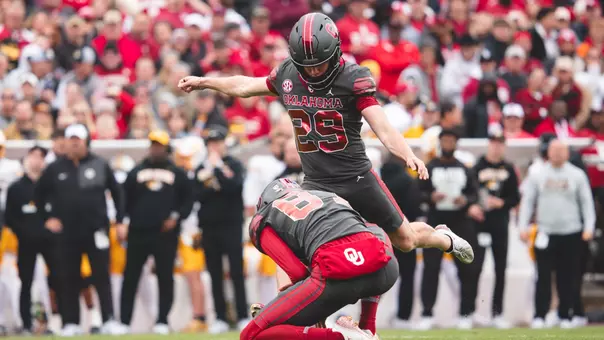 Tate Sandell and holder Jacob Ulrich in OU's dark gray Unity uniforms, showing Sandell kicking a field goal at Gaylord Family - Oklahoma Memorial Stadium.